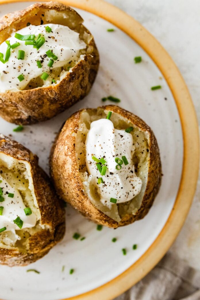 Three baked potatoes split open on a plate with sour cream, fresh chives, and seasoning.