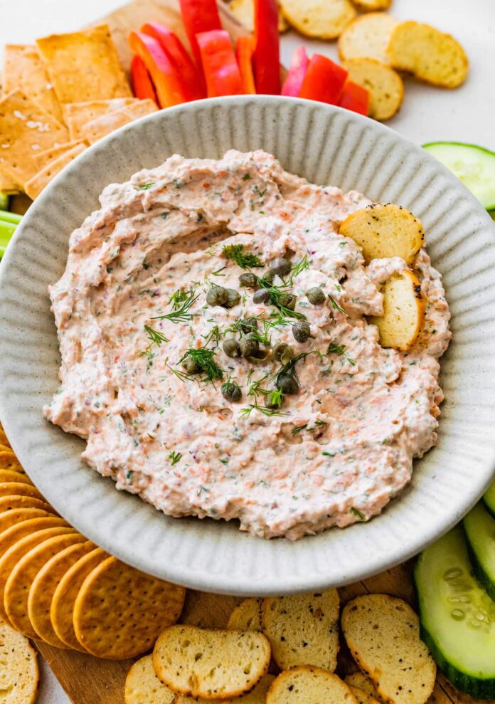 A bowl of smoked salmon dip, garnished with fresh dill and capers, served with crackers and fresh vegetables.