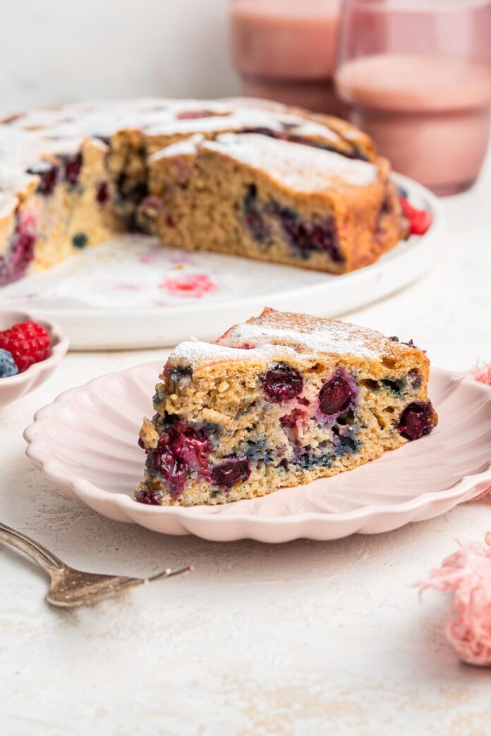 A slice of mixed berry cake on a light pink plate. The entire berry cake is in the background on a white plate.