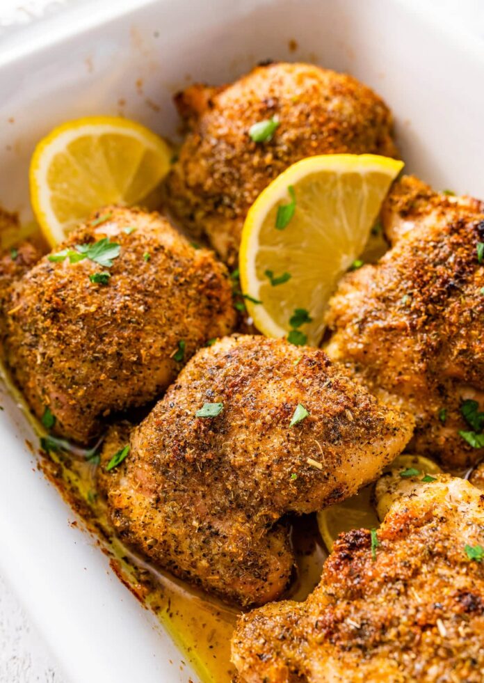 Six baked Greek chicken thighs in a white baking dish. The chicken is topped with fresh parsley and lemon slices.