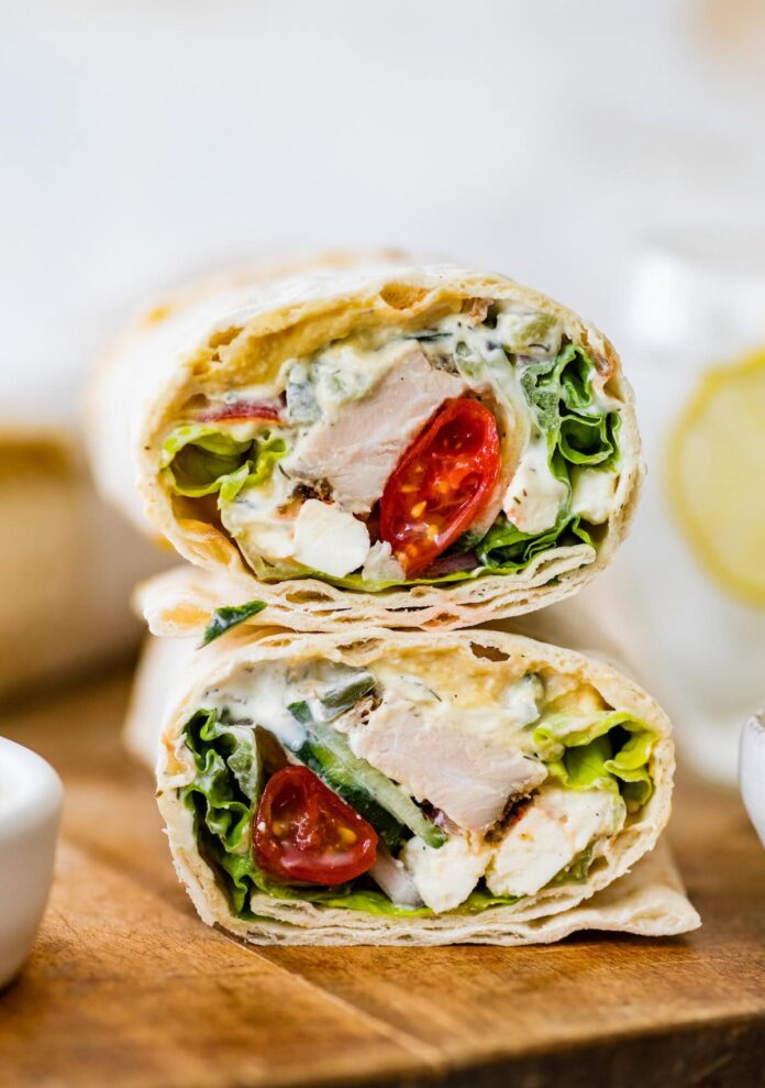 Two halves of a Greek chicken wrap stacked on top of each other on a wooden cutting board. You can see the tomatoes, chicken, lettuce and sauce inside the wrap.