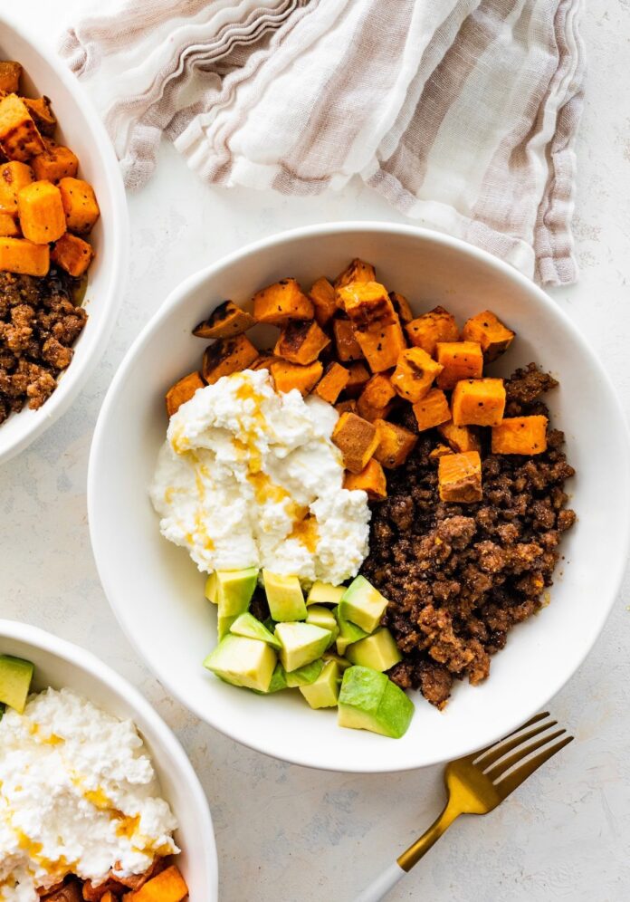 Ground beef, roasted sweet potatoes, cottage cheese and chopped avocado in a white bowl topped with a drizzle of honey.